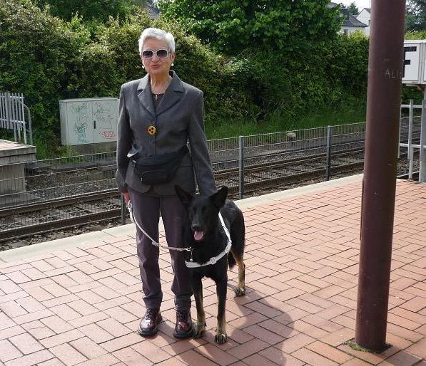 Dieses Bild zeigt Irma Schulze-Schlüter. Zu Ihrer linken steht Anna, eine schwarze Schäferhündin. Sie stehen beide am Bahnsteig in Sankt Augustin.